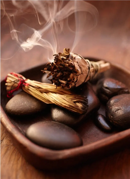 Smudge bundle in a bowl on rocks with smoke rising, used for cleansing and spiritual rituals.
