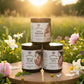 Three jars of 'Divine Beginnings' product on a wooden stand with a natural background of flowers and greenery.