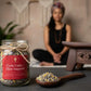 Jar labeled 'Cycle Calm + Flow Support' with a wooden spoon of herbs on a table, blurred figure in the background.