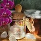 A beautiful glass jar of the Rebirth All-Natural Organic Crystal Bath Soak next to a wooden spoon beside an orchid in the sunlight. 